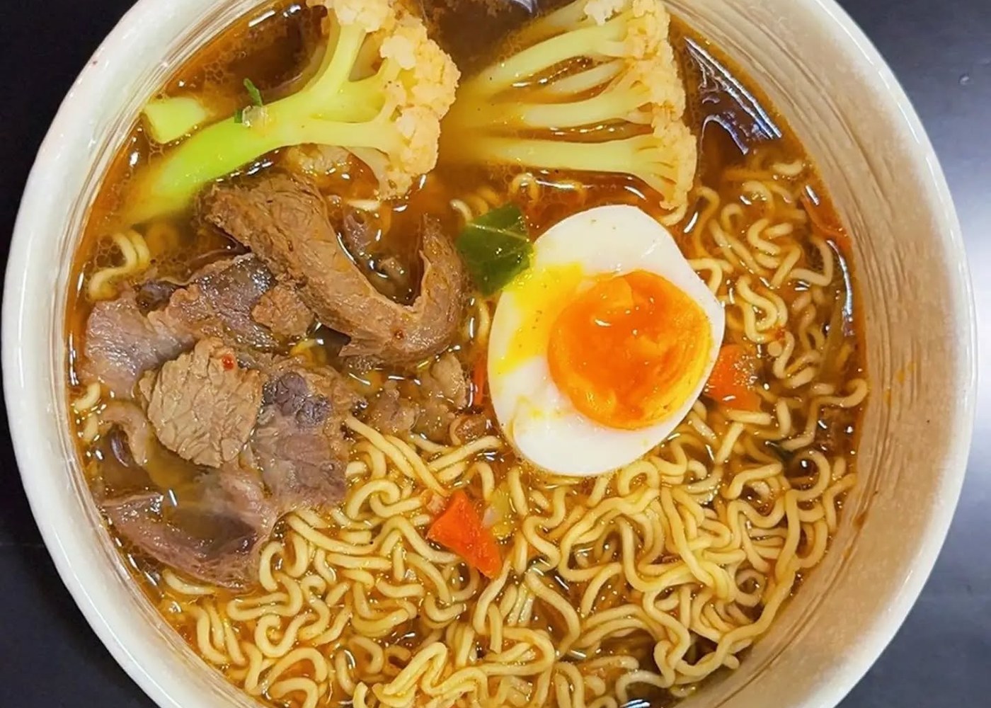 Ciotola bianca di ramen in brodo ambrato con fettine di manzo, mezzo uovo morbido, cavolfiore e carota