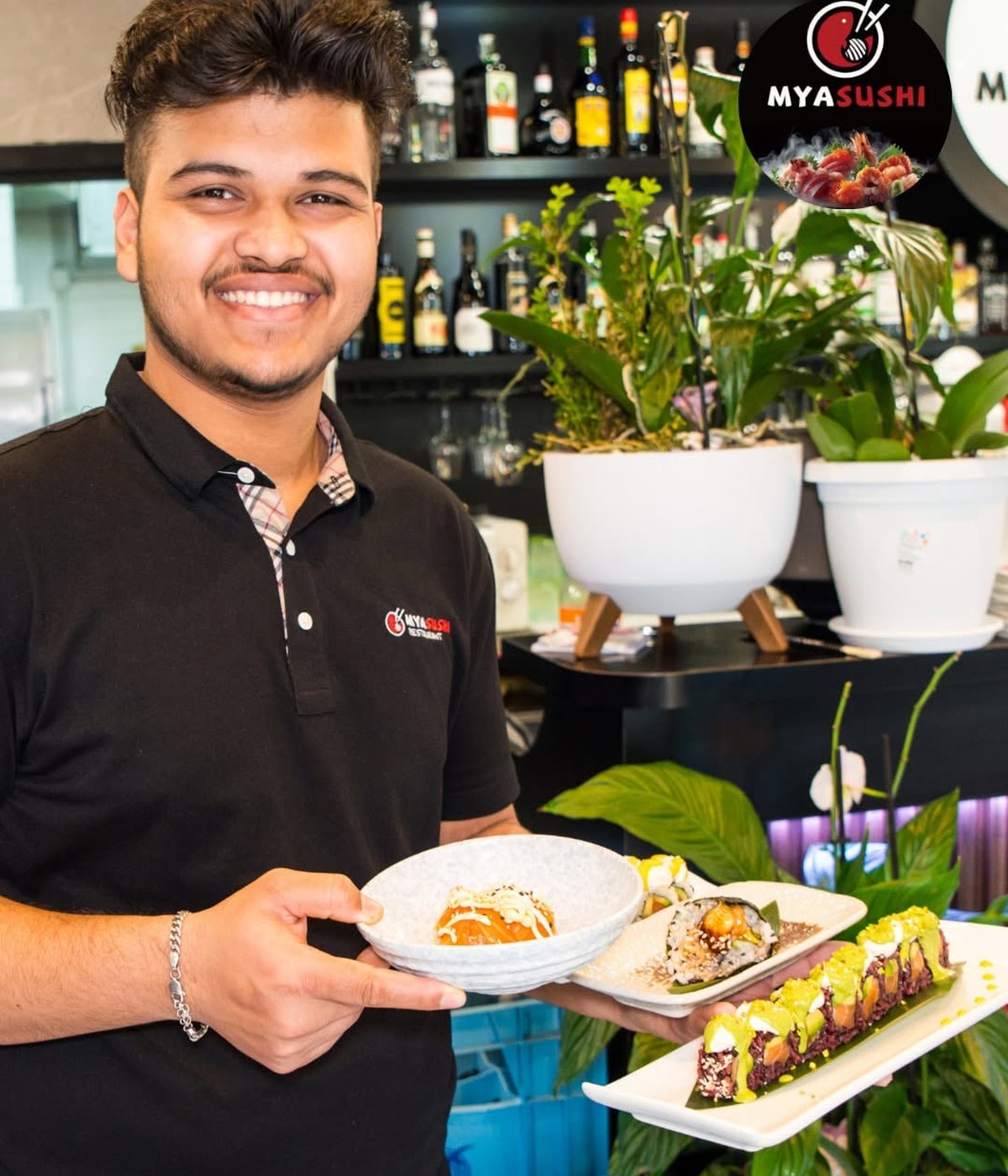 Cameriere sorridente con polo nera Mya Sushi regge tre piatti di sushi roll davanti a bancone bar con bottiglie e piante
